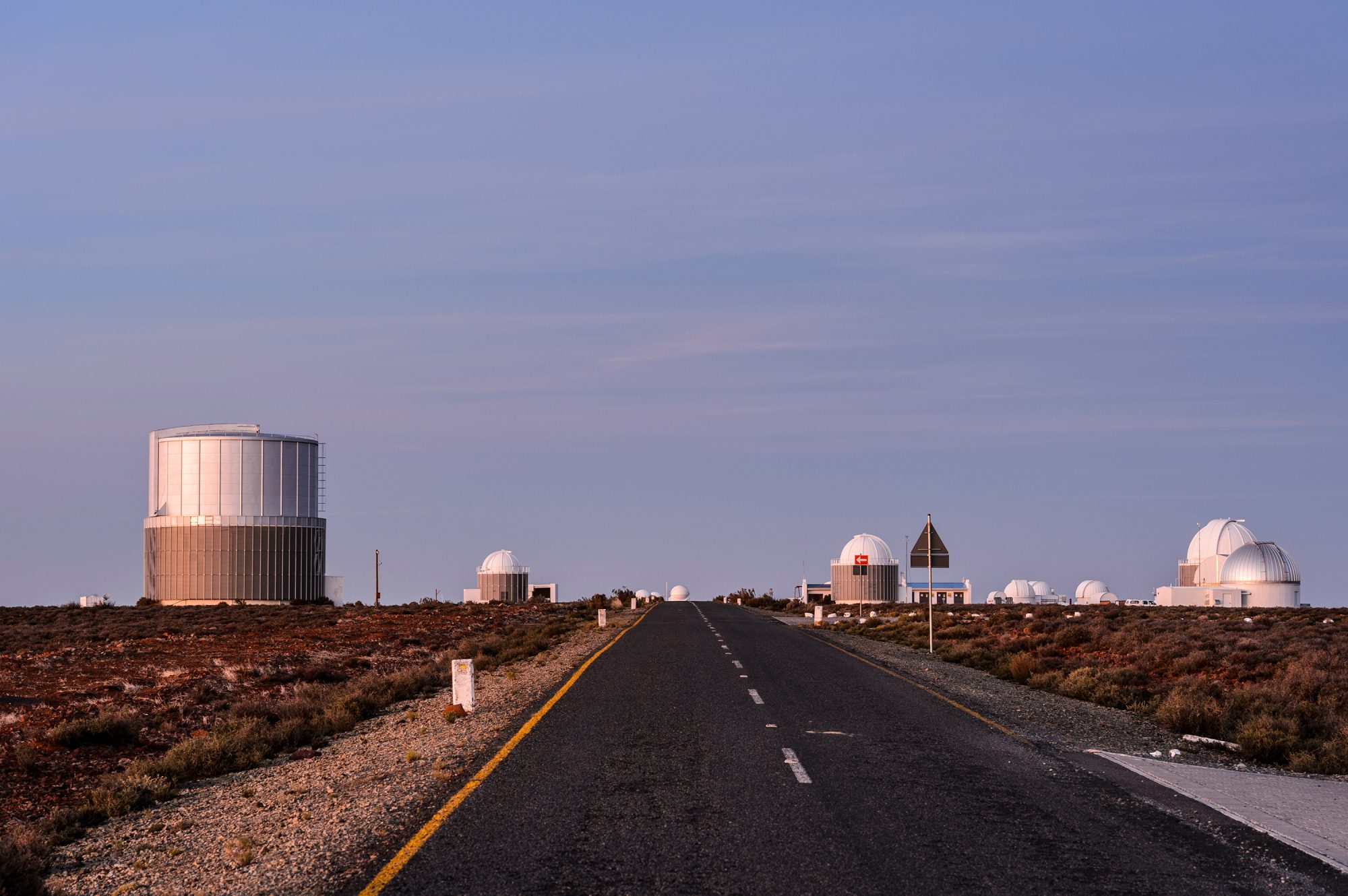 The paved road to the Southern African Large Telescope, showing smaller dome shaped structures and little vegetation around them. Big blue sky.