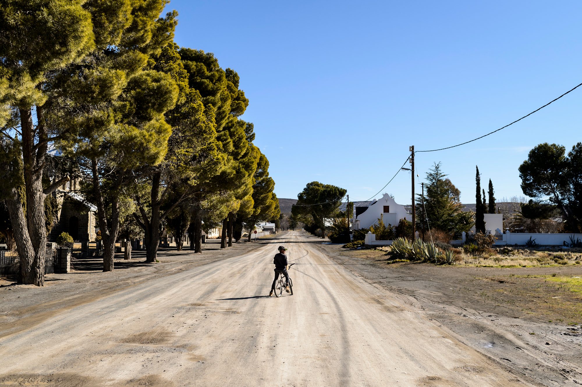 A boy pauses on his bicycle on a dusty rode in Sutherland, South Africa. Trees, a few houses, blue sky.