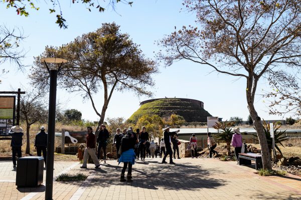 Exterior view of the Maropeng Museum in South Africa, a mounded structure in the background, visitors milling in the foreground on a bright day.