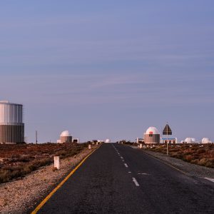 The paved road to the Southern African Large Telescope, showing smaller dome shaped structures and little vegetation around them. Big blue sky.