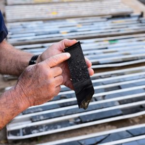 Nic Beukes's hands point to a dark black core sample, trays of samples are out of focus but visible in the background.