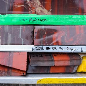 Trays of rock core samples aligned and labeled, showing depth. The samples are striated in grays, oranges, deep oranges.
