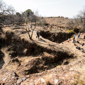 Looking down at the archaeological site from a hilltop, a few people can be seen walking around the site on a bright day.