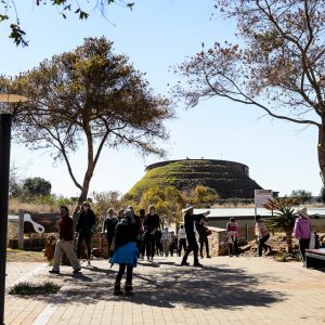 Exterior view of the Maropeng Museum in South Africa, a mounded structure in the background, visitors milling in the foreground on a bright day.