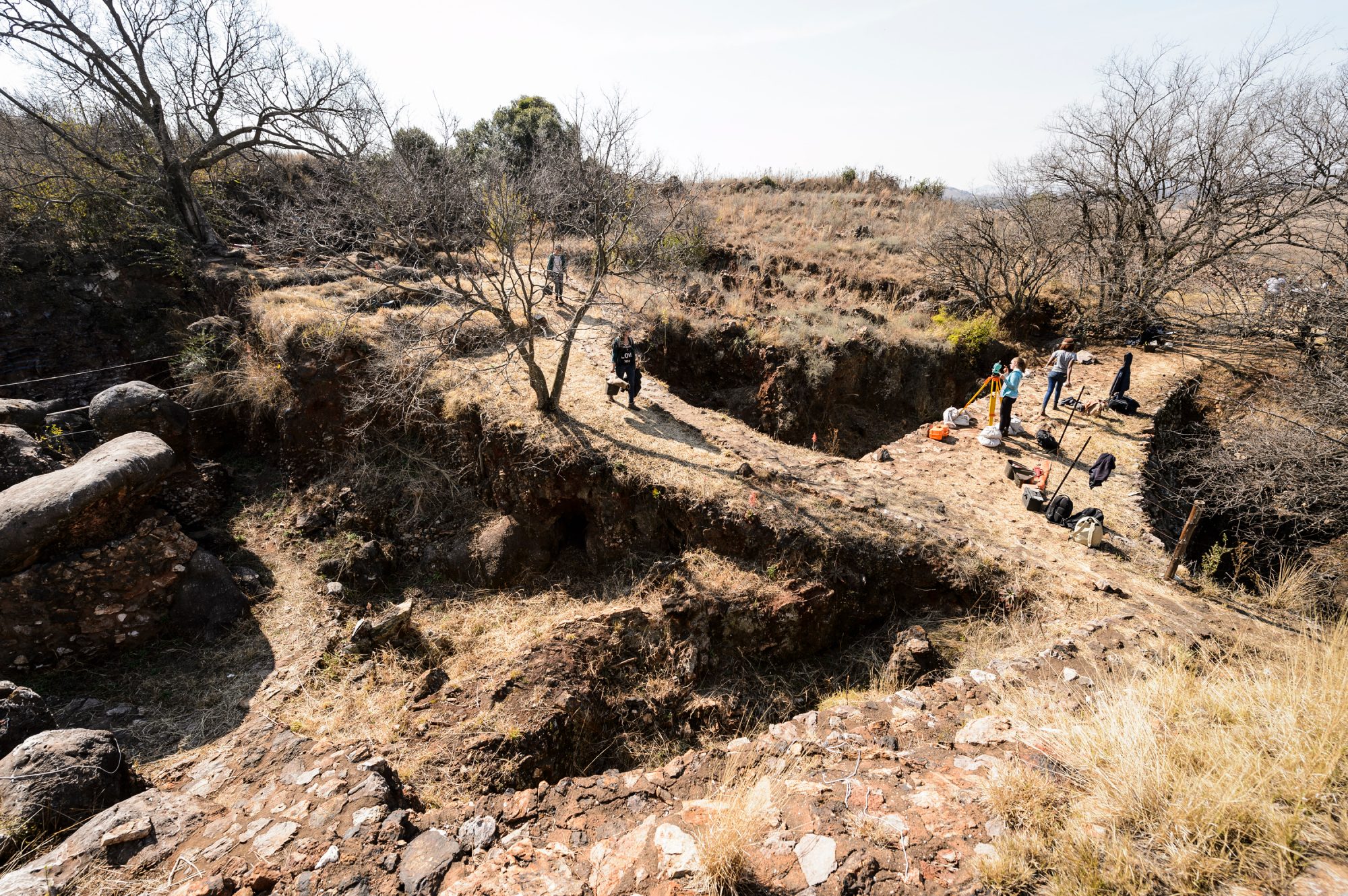 Looking down at the archaeological site from a hilltop, a few people can be seen walking around the site on a bright day.