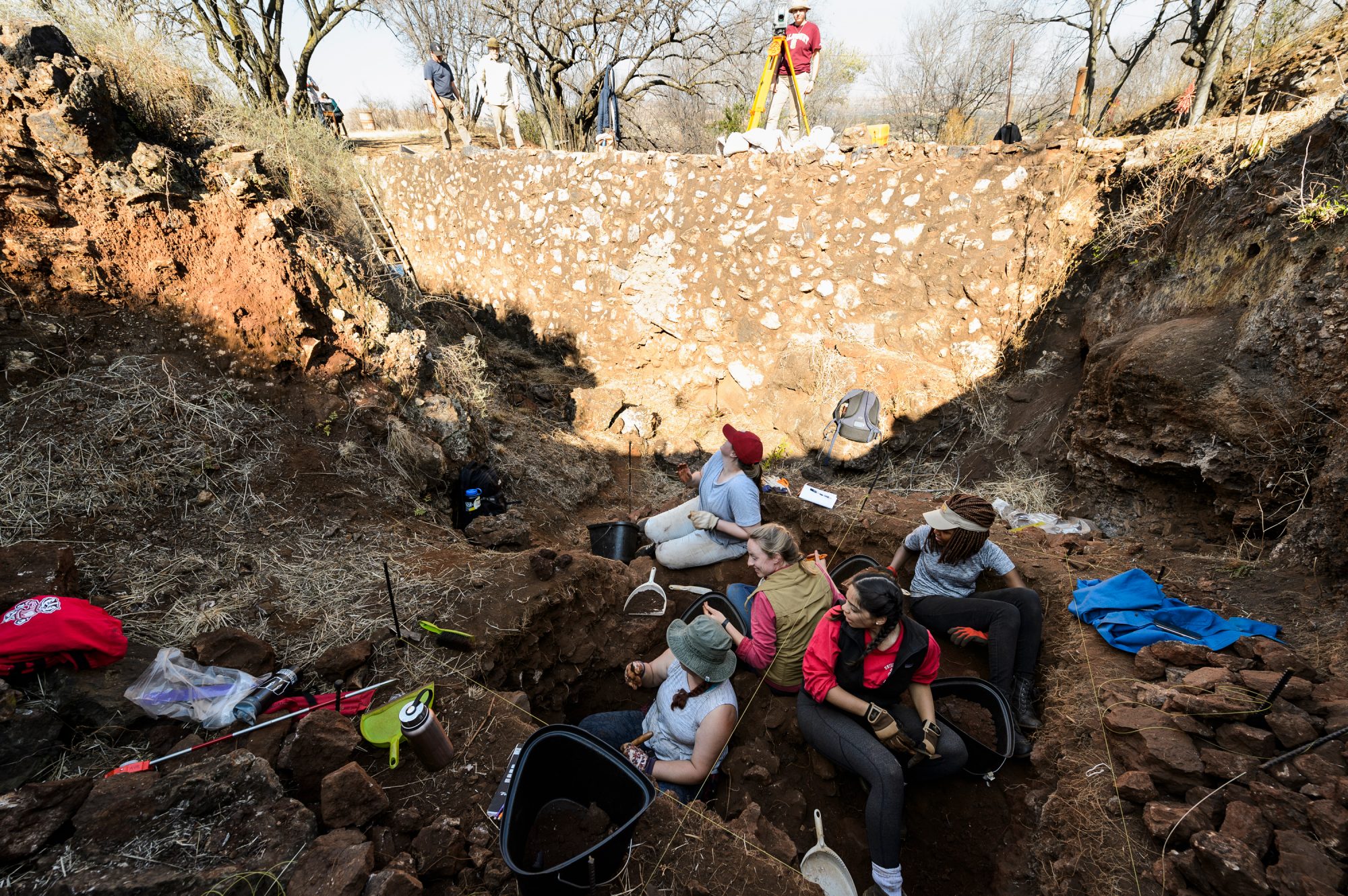 Five field school students at the bottom of a shallow archaeological site digging on a bright day.